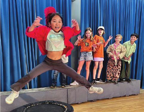 Lexington School students rehearsing Here Be Dragons on stage, one leaping mid-air